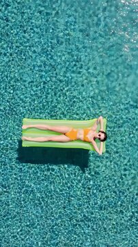 Aerial view of a young woman wearing an orange bikini and sunglasses relaxing on a green inflatable mattress in a turquoise swimming pool during a sunny day. Vertical Shot