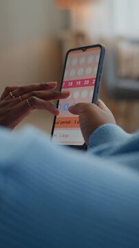 Vertical shot of unrecognizable woman tracking menstrual cycle in app on smartphone while sitting at home