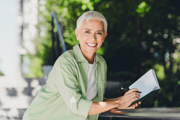 Confident elderly woman enjoying the outdoors writing in a notebook in a casual green shirt with...