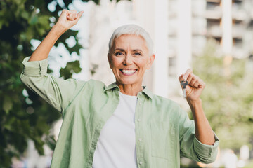 Obraz premium Confident senior woman in a casual green shirt celebrates outdoors with keys in hand on a sunny city street in a modern urban neighborhood