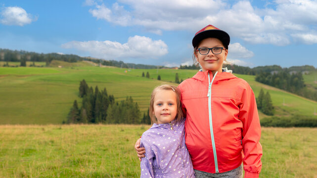 Two sisters hug in mountain meadow landscape, smiling kids in colorful jackets and cap with scenic countryside and cloudy blue sky, family outdoor travel portrait.
