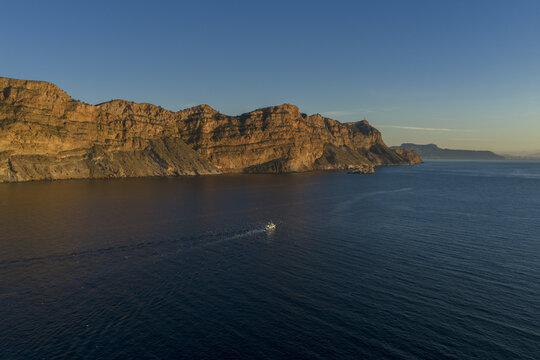 Aerial view of the rugged cliffs bathed in the warm glow of the setting sun meet the dark blue sea, a lone boat cutting through the tranquil waters, Ocean, Alicante, Spain.
