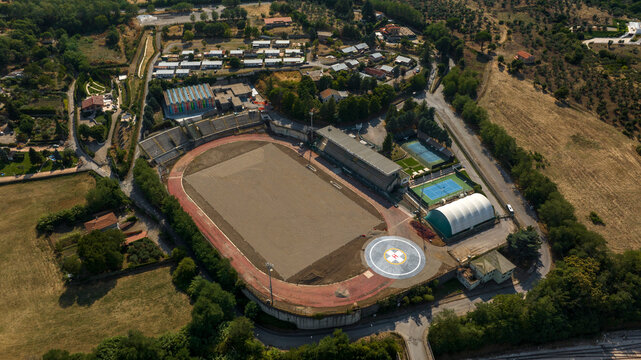 Aerial view shows a sports complex featuring a dirt athletics track and field, tennis courts, and a large indoor tent. Surrounded by lush green trees and a small residential area.