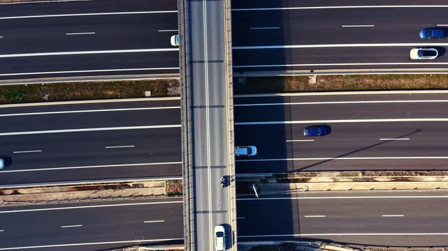 Vehicles move on multiple lanes below a bridge. The scene captures an urban area during daylight. Shadows from the bridge stretch over the road surface.