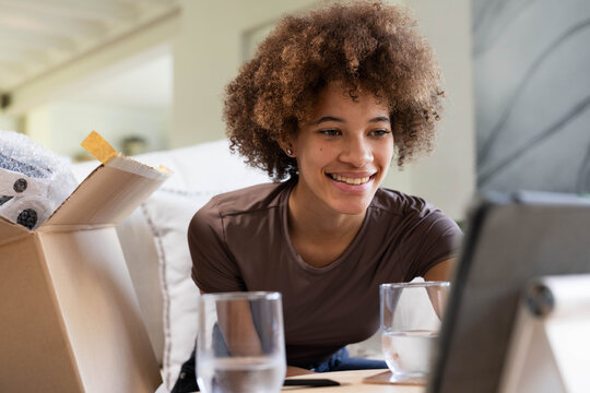 African American woman sitting on couch in living room, watching tablet with open box and glasses