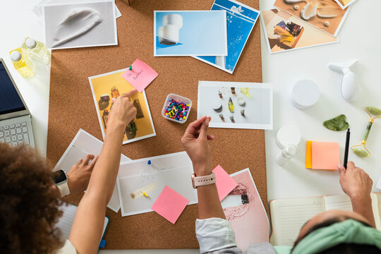 Printed product layouts are being arranged and pinned on corkboard at white tabletop with pushpins