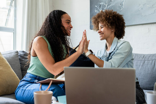 Two female friends high-fiving while sitting on gray sofa at home with laptop and notebook