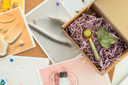 Small cardboard gift box is resting on corkboard with jade roller, gua sha, pink filler