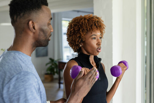 African American man coaching woman in black top doing bicep curls at home with purple dumbbell