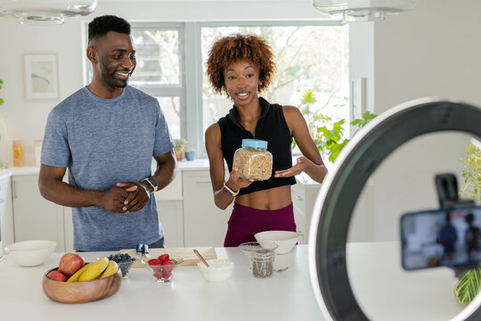Diverse couple presenting breakfast demonstration in bright home kitchen, holding jar of oats