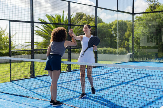 Two female paddle players exchanging high fives at padel net wearing white dress and holding paddle