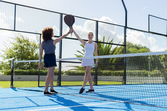 Female friends wearing white tennis dress and navy skirt raising padel rackets at blue court net