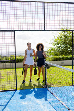 Diverse female friends standing at open metal gate on blue court holding paddles and bottle