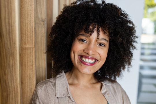 African woman smiling and leaning on wooden slat wall, wearing beige shirt and green top