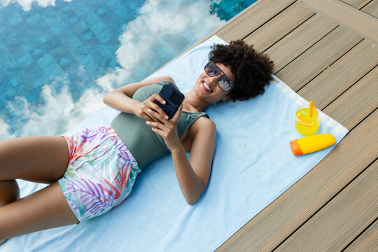 African woman reclining on blue towel by pool, wearing olive-green top and holding smartphone
