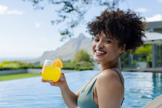 African American woman standing at pool edge holding glass with orange wearing swimsuit, copy space