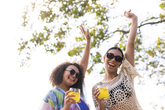 African female friends holding yellow drinks, wearing sunglasses and crochet coverup under trees