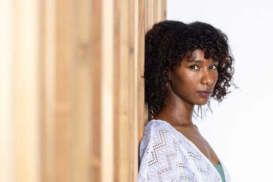 African American woman leaning against studio wood slats, in white knit, green camisole, copy space