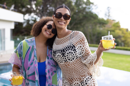 African American women standing poolside at home in swimsuits and sunglasses, holding orange drinks