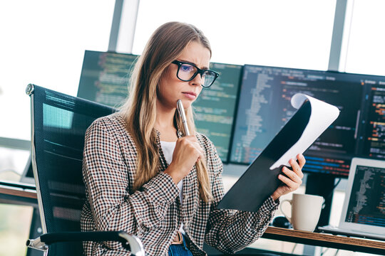 Young female programmer reviewing code on printout while thinking at a modern office desk with multiple monitors and laptop