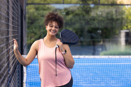 African American woman holding black paddle on court by fence, net, wearing pink top, smartwatch