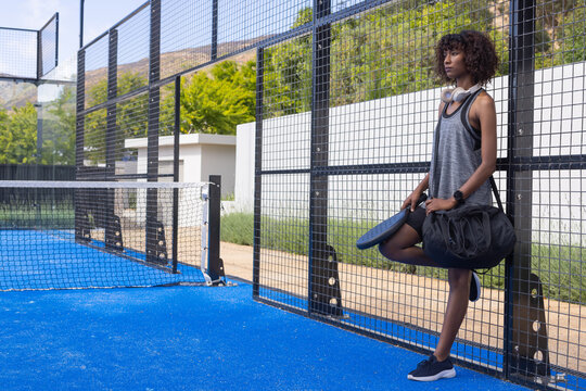 African woman leaning against mesh fence on blue paddle court holding paddle and duffel, copy space