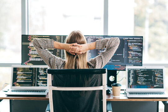 Relaxed software developer leaning back at a multi‑monitor coding workspace with laptop screens and coffee in a bright modern office