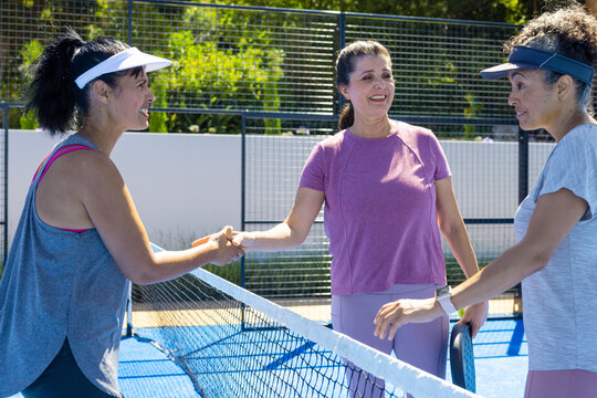 Diverse women in sportswear shaking hands at net on blue paddle court with paddle and visors