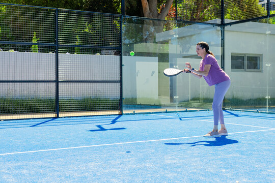 Woman standing on blue court holding white padel racket watching green ball near glass, copy space