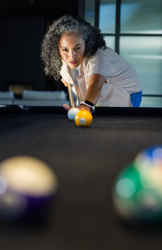Senior African American woman leaning and aiming cue stick toward yellow ball on dark pool table