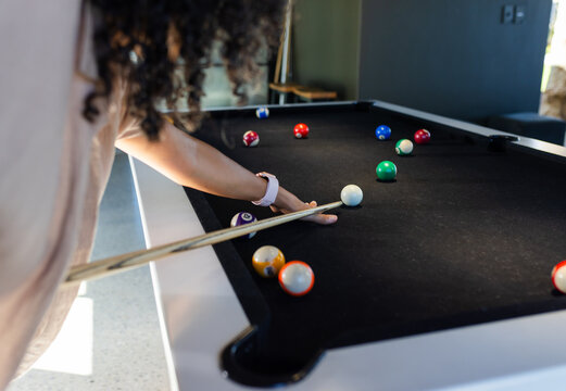 African American woman leaning on black-felt pool table at lounge aiming cue at cueball with watch