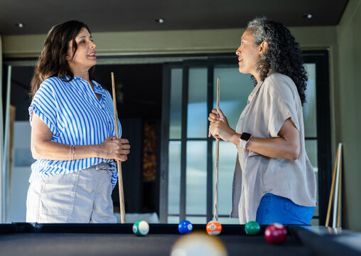 Diverse female friends standing, chatting at home rec room pool table holding cues, billiard balls