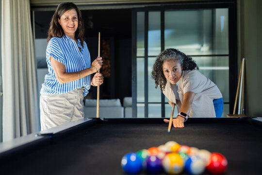 Diverse female friends lining up shot and holding cues at black-felt pool table with racked balls