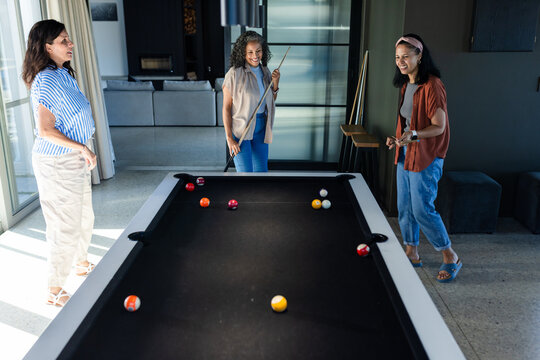 Diverse female friends playing pool in home lounge at black-felt table with billiard balls and cues