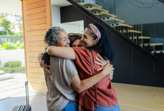 Three women including senior hugging and smiling in home entry with sunglasses and rolling suitcase