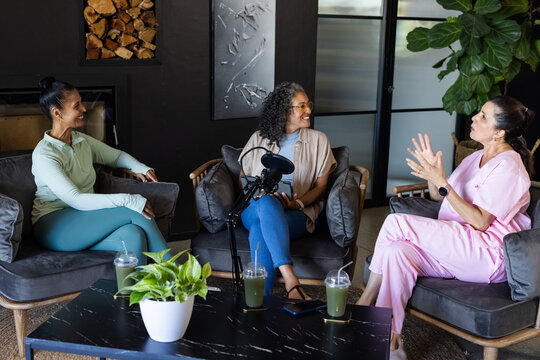 Diverse female colleagues discussing interview in lounge wearing scrubs with microphone and drinks
