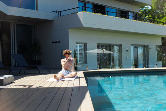 Man sitting cross-legged on wood pool deck white towel wearing trunks and shades sipping from glass