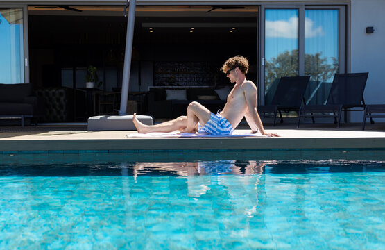 Man sitting on towel at pool edge wearing striped trunks on sunlit deck by sliding doors