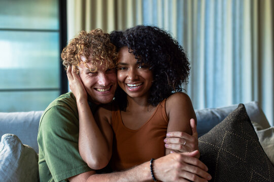 Couple sitting on sofa by window, smiling, wearing green tee and rust tank, showing ring