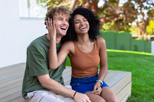 Couple with African American woman and man laughing, sitting on low wooden porch showing wristwatch