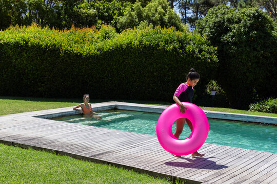 Diverse female pair teenage girl walking deck holding pink ring and woman sitting pool, copy space