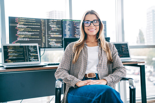 Smiling female software developer at desk with multiple monitors displaying code in a bright modern office workspace