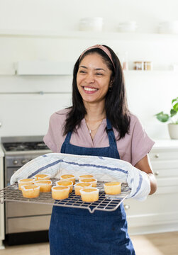 African woman smiling holding wire rack of dozen cupcakes wearing denim apron with towel in kitchen