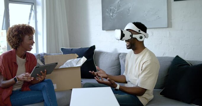 Diverse couple unpacking VR headset on gray sofa, woman guiding tablet while man testing VR world