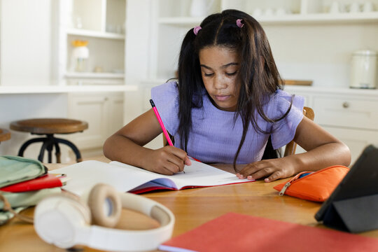 School-age girl writing in open notebook at kitchen table with pink pencil, headphones, backpack