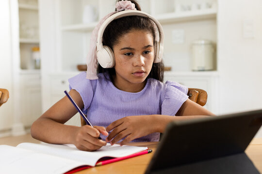 African American child sitting at table with headset writing in notebook with pencil viewing tablet