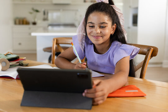 African American preteen girl sitting at home dining table using tablet with pencil near notebook