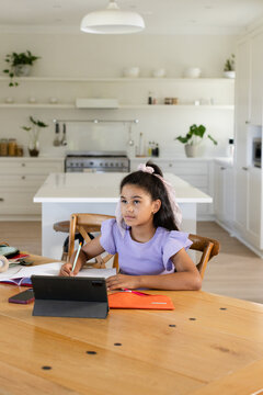 African American preteen girl sitting at kitchen table using tablet with stylus and writing notes