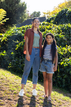 African woman and youth girl standing on sunlit grassy slope in yard wearing denim, sandals