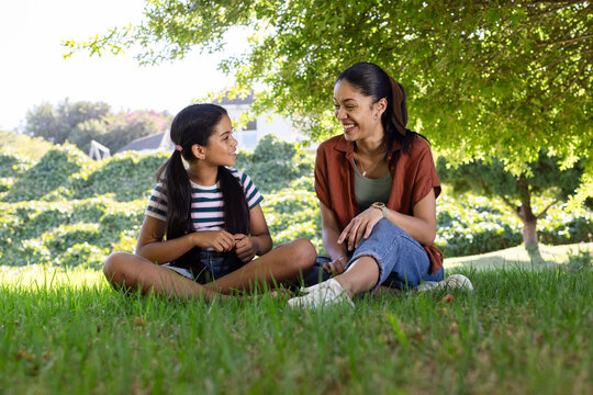 African American mother and child sitting on grass under large shade tree, wearing white sneakers